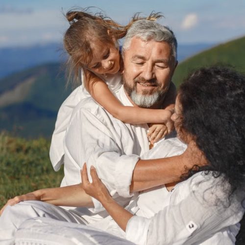 A joyful family moment outdoors, showing grandparents hugging their granddaughter under a sunny summer sky.